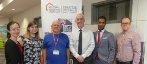 Nic stands in front of a "National Housing Strategy" banner with CMHC representatives.