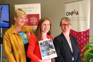 Three women smile, holding up the Affordable Housing Plan for Ontario, in front of banners reading "CHF Canada" and "ONPHA"