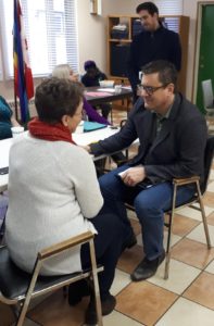 Sitting facing one another in chairs, a man in a blazer speaks with a woman in a white sweater