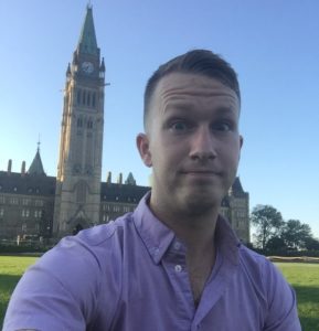 A white man with short hair and a pink button-up shirt poses in front of the Parliament of Canada