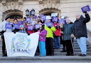 A group of co-op members, adults and children, stand on the steps of the Legislature holding a banner and purple #IncludeCoop signs