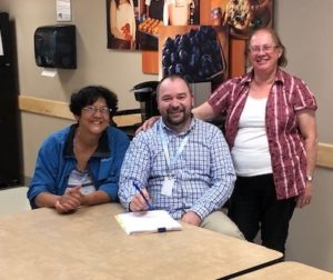 Three people sit and stand proudly around a table, about to sign paperwork