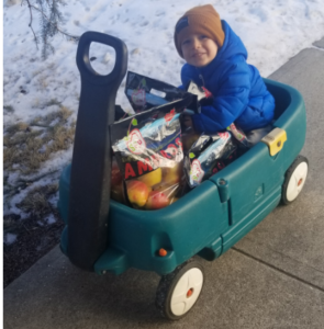 A small child smiles from a wagon filled with bags of apples