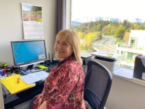 A person with blond hair and a colourful red shirt smiles at their desk, with a beautiful view of trees out the window