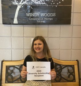 A person with long blonde hair sits on a bench under a sign reading Windy Woods Co-op, holding up a sign that says "CHF Canada 2020 Diversity Scholarship recipient"