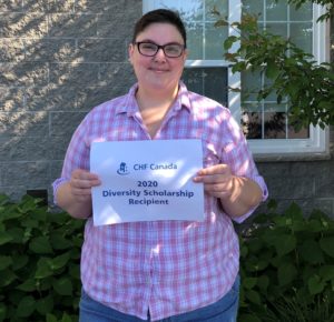 A person with glasses and a checked shirt stands in a garden holding up a sign that says "CHF Canada 2020 Diversity Scholarship recipient"