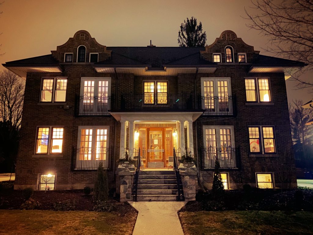 A stately low-rise brick apartment building is brightly illuminated against an evening sky