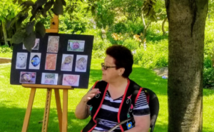 A woman with short dark hair and glasses stands beside a selection of art pieces on an easel