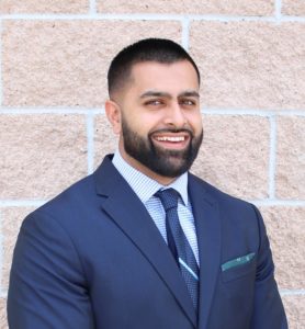 Irfaan, who has shortly clipped hair and beard, stands in front of a brick wall smiling and wearing a blue suit