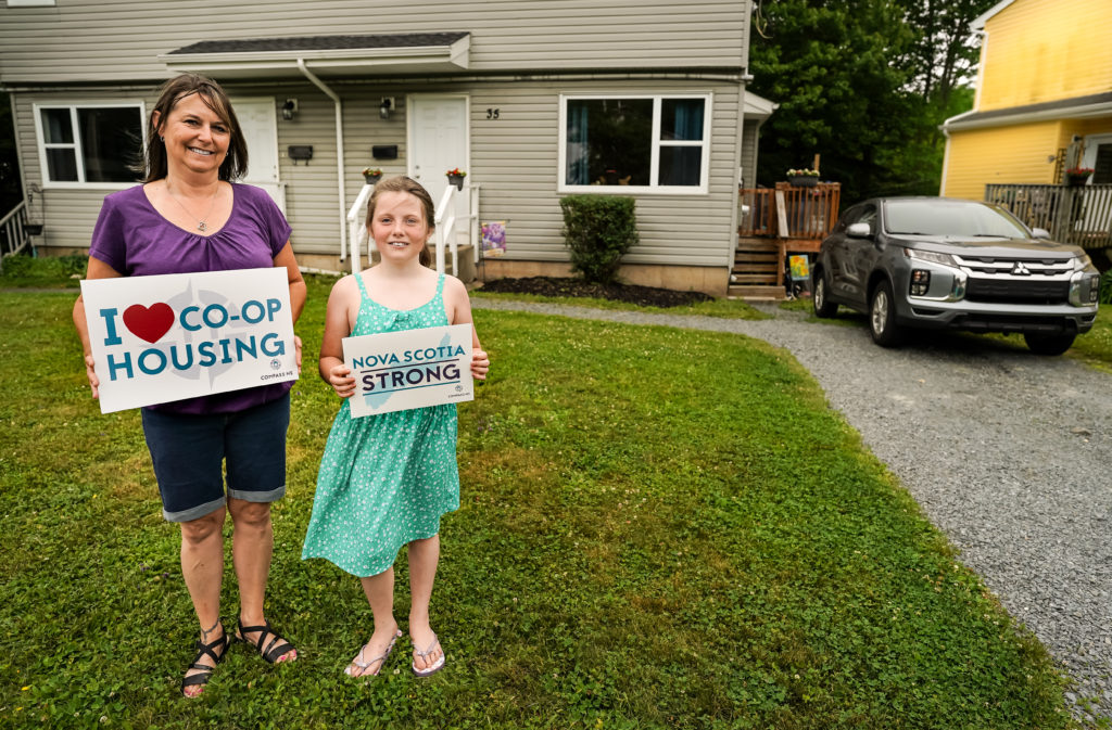 A mother and daughter stand smiling on the lawn of their co-op home. They hold signs saying "I love co-op housing" and "Nova Scotia Strong"."