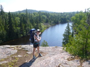 Cynthia stands on a cliff in hiking gear, gesturing out over a lake and trees