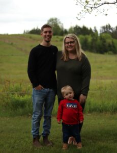 A young man and woman in a long field stand behind their son, a toddler.