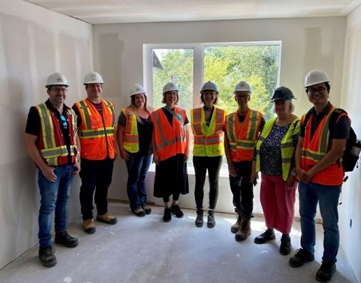 A group of men and women in construction hats and safety vests pose in a room under construction.