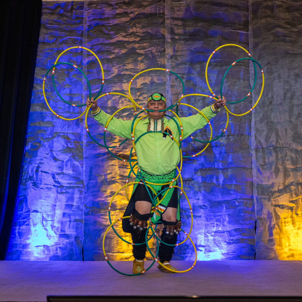 A person in a green outfit performs on stage with multiple colorful hoops arranged around their body, set against a textured backdrop with blue and yellow lighting—highlighting the spirit of co-operative housing at a CHF Canada event.