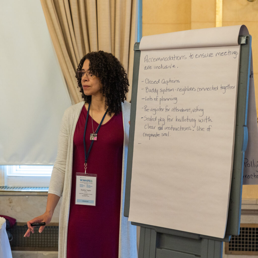 A woman stands next to a flip chart listing accommodations for inclusive meetings in a conference room, holding a marker and wearing a name badge, as part of a CHF Canada co-operative housing workshop.
