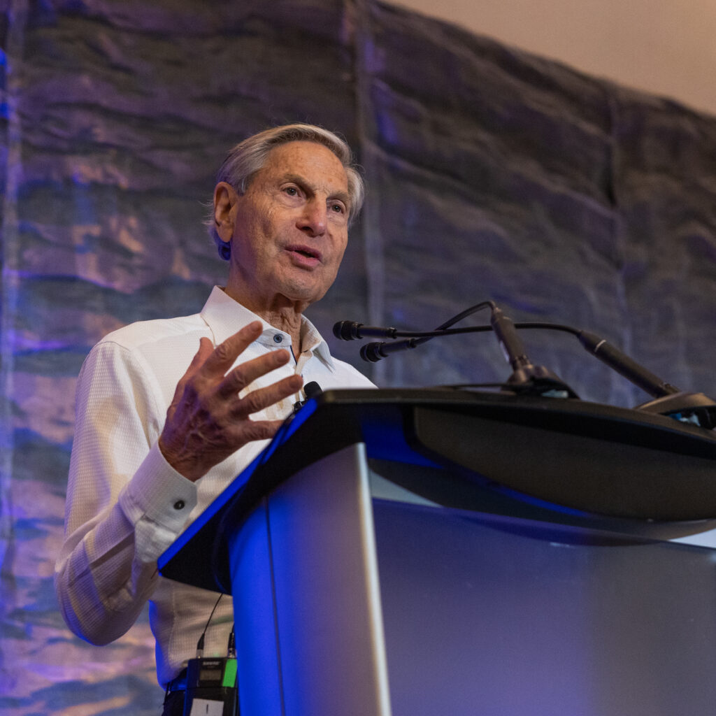 An older man in a white shirt speaks at a podium with microphones, addressing issues of Co-operative Housing, standing in front of a textured, grayish-blue background.