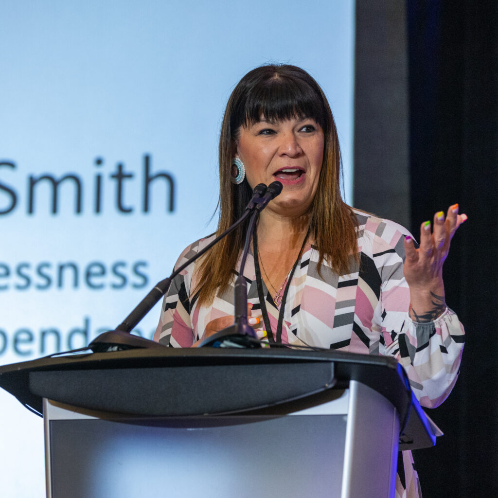 A woman stands at a podium speaking into two microphones, gesturing with one hand, as a presentation screen in the background displays partial text about Co-operative Housing and CHF Canada.