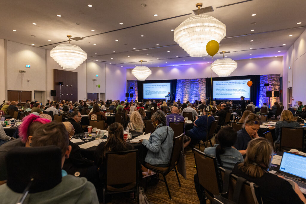 Large conference room filled with attendees seated at tables, facing two large screens displaying CHF Canada presentation slides on co-operative housing at the front of the room.