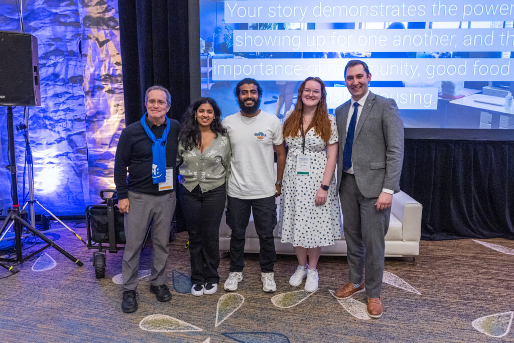 Five people pose together in front of a stage with a large screen displaying text about Co-operative Housing, standing on a carpeted floor with lighting equipment visible at the CHF Canada event.