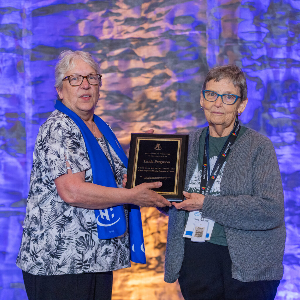 Two older women stand in front of a blue-lit stone wall, holding a plaque together. One wears a blue scarf; the other glasses and a lanyard, celebrating their achievements in Co-operative Housing with CHF Canada.