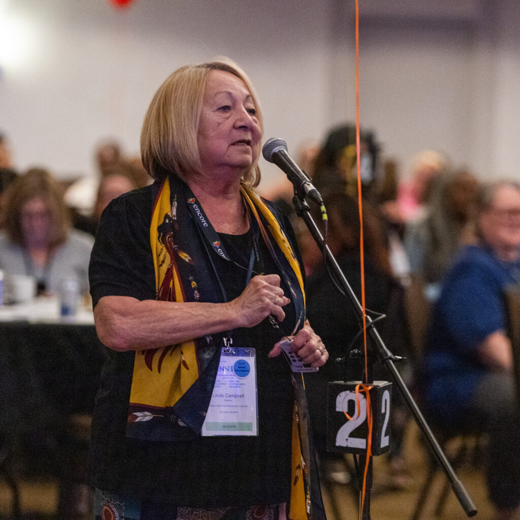 A woman speaks into a microphone at a CHF Canada conference, wearing a name badge and scarf, with an audience seated in the background, discussing co-operative housing initiatives.
