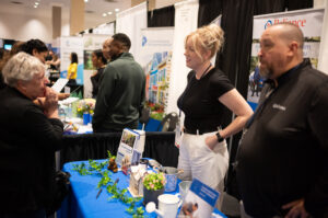 Two people stand behind a booth at an indoor event, talking to a visitor across the table decorated with brochures and small plants, promoting CHF Canada and the benefits of Co-operative Housing.