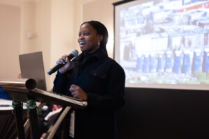 A woman speaks into a microphone at a podium, with a laptop and a projector screen displaying images about Co-operative Housing in the background at a CHF Canada event.