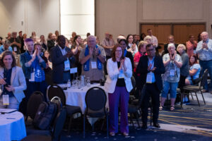 A group of people stand and clap in a conference room at a CHF Canada event, some wearing lanyards, with round tables and empty chairs in the foreground, celebrating co-operative housing achievements.
