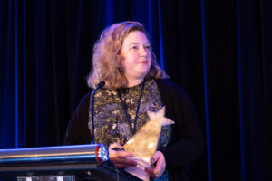 A woman stands at a podium holding a star-shaped award, with a blue curtain backdrop, celebrating achievements in co-operative housing at a CHF Canada event.