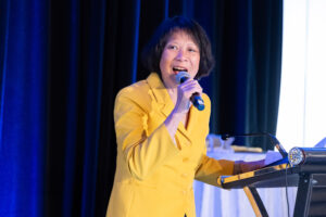 A woman in a yellow blazer speaks into a microphone while standing at a podium, addressing the audience about co-operative housing at a CHF Canada event.