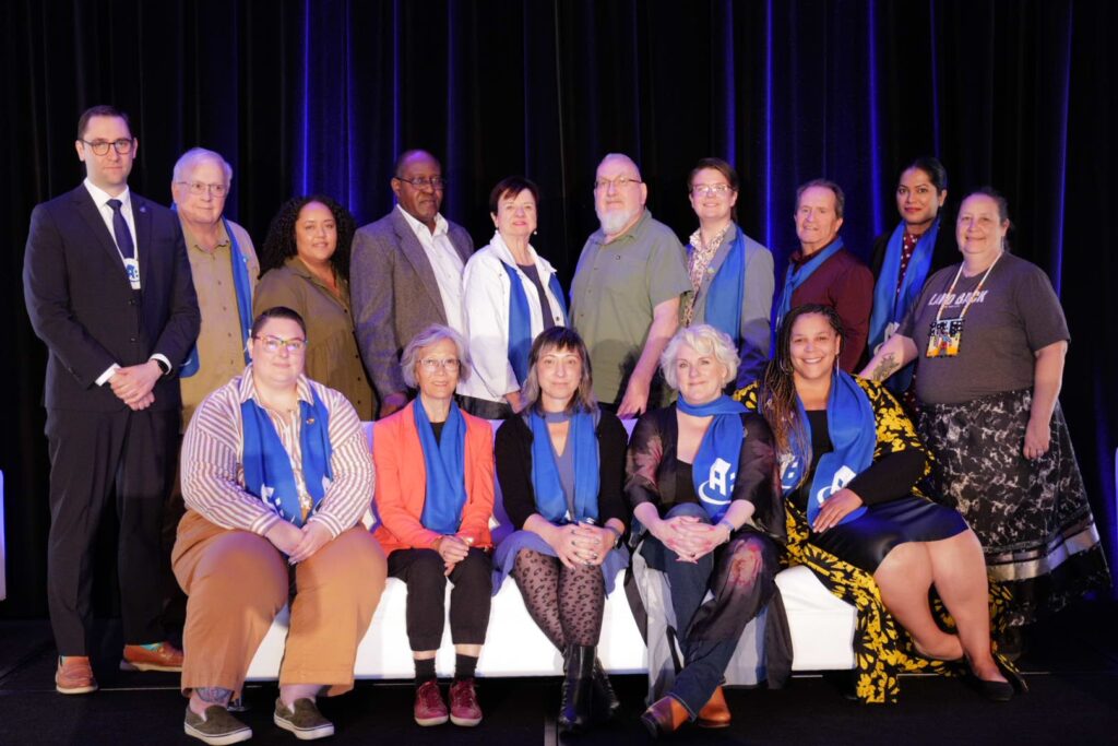 A group of fifteen people pose together indoors, some seated and some standing, wearing blue scarves or accents in front of a dark curtain, representing their commitment to co-operative housing and the values championed by CHF Canada.