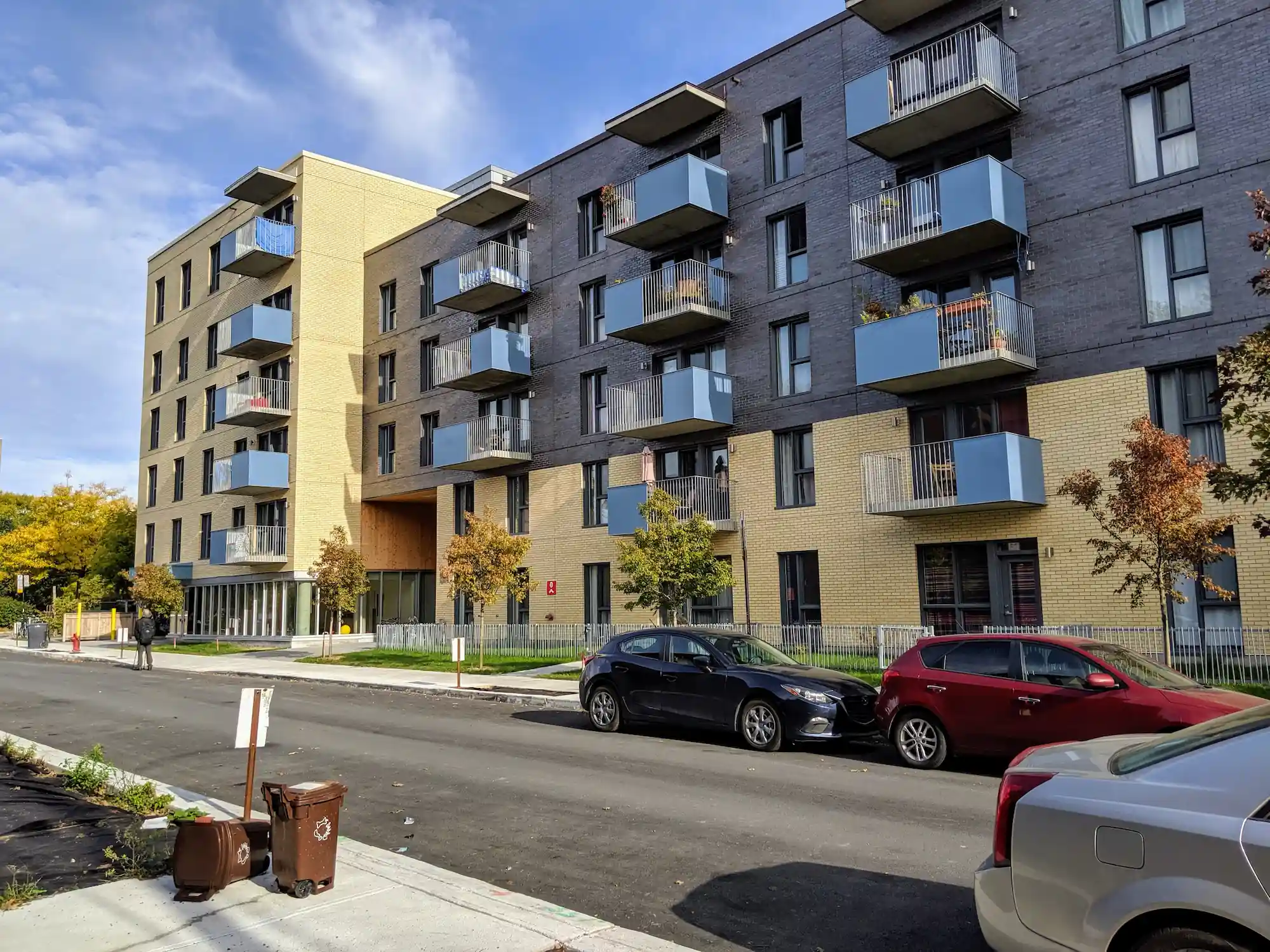 A modern mid-rise apartment building with balconies, parked cars along the street, and small trees lining the sidewalk on a sunny day, showcasing the appeal of co-operative housing supported by CHF Canada.