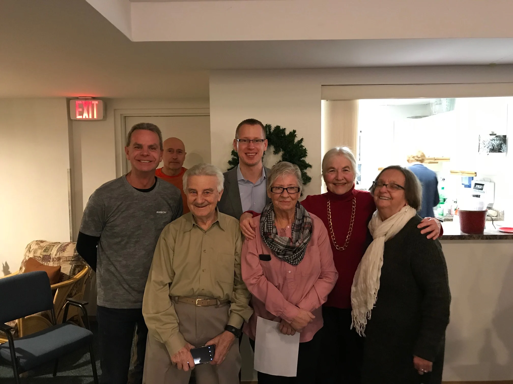 Seven adults pose and smile for a group photo indoors, with a kitchen area and chairs visible in the background, capturing a lively moment at a CHF Canada Co-operative Housing gathering.