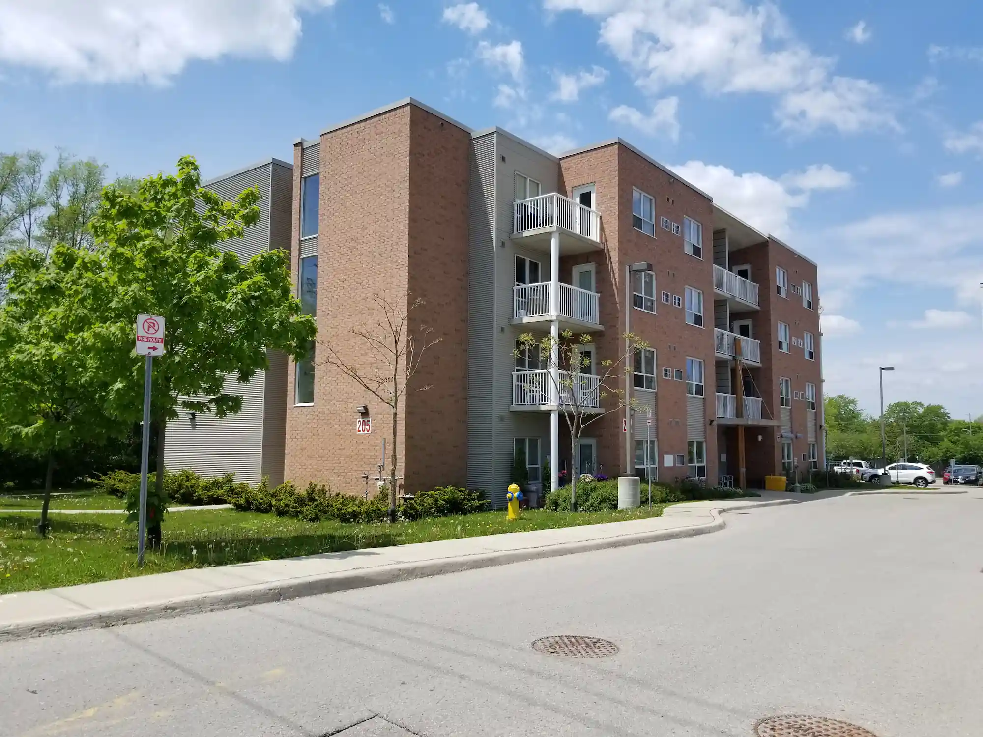 A three-story brick and siding co-operative housing apartment building with balconies, surrounded by greenery under a partly cloudy sky. A no parking sign and fire hydrant are visible near the entrance.