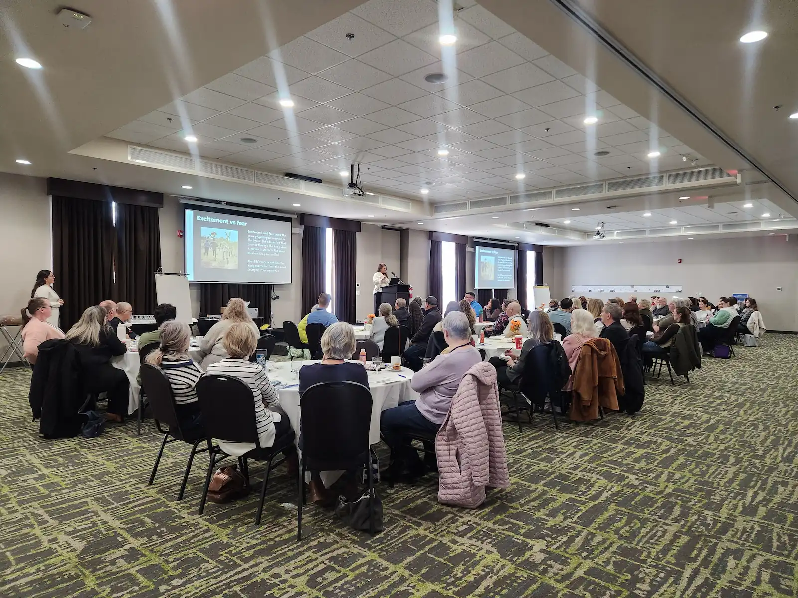 A group of people sit at round tables in a conference room, watching a presentation on co-operative housing projected onto a screen at the front, likely during a CHF Canada event.