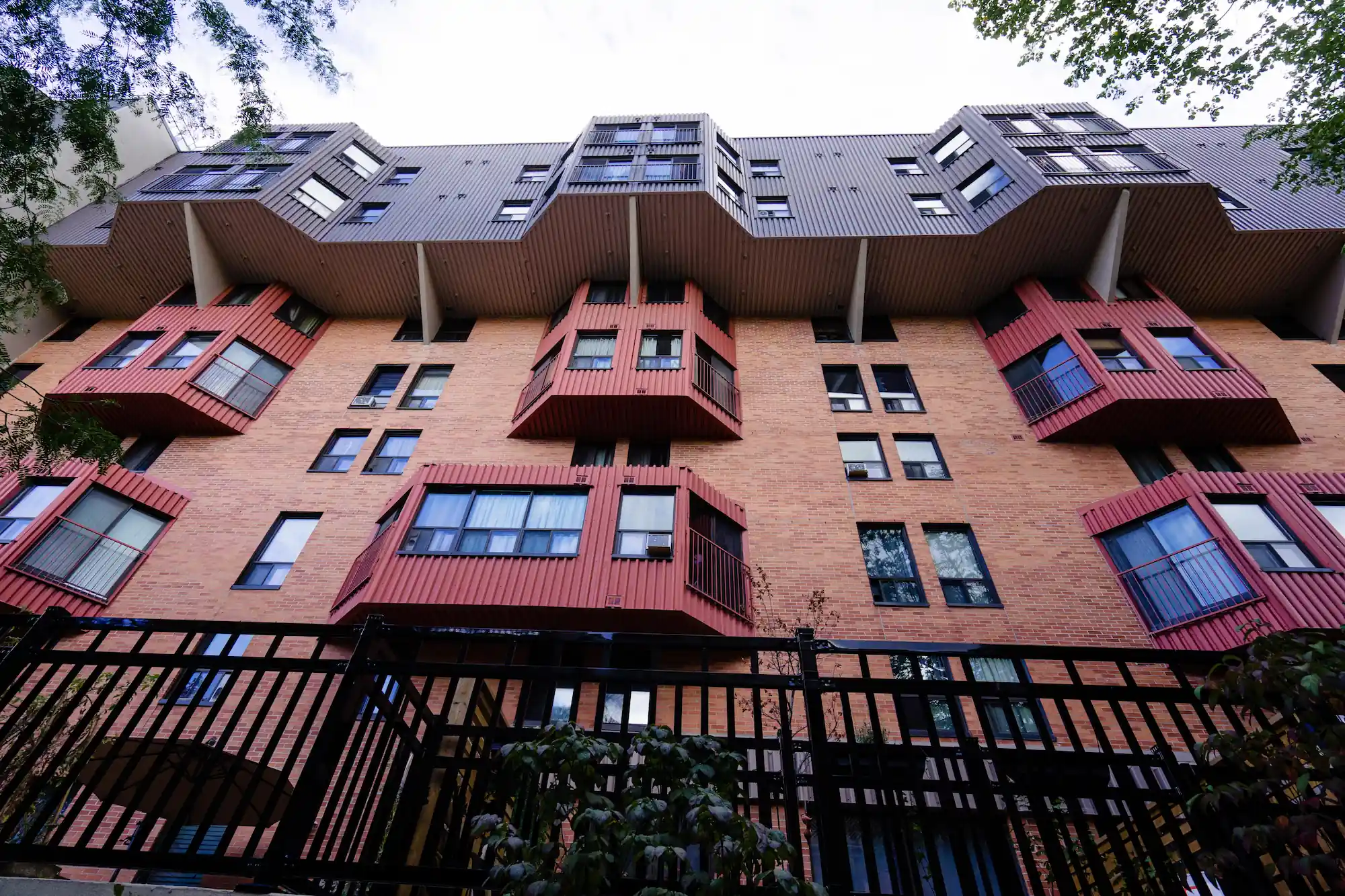 A multi-story brick co-operative housing apartment building with protruding red balconies and a dark upper level, viewed from below behind a black metal fence.