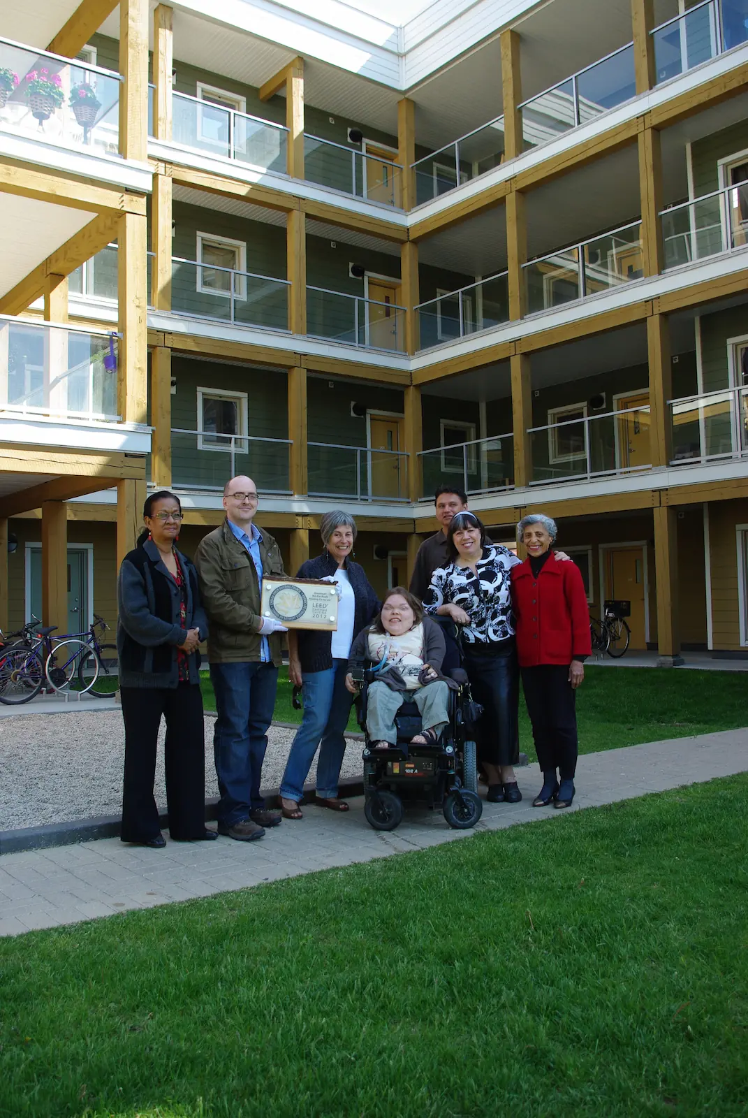 A group of seven people, including one in a wheelchair, pose and smile in front of a modern, multi-story co-operative housing apartment building.