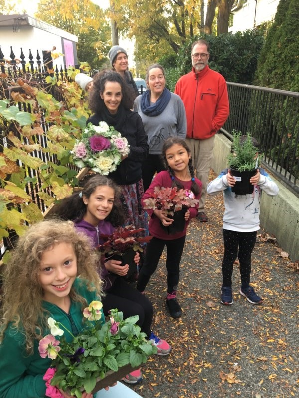 A group of five children and four adults stand outdoors on a sidewalk, smiling and holding potted plants and flowers, celebrating community spirit in co-operative housing with autumn foliage in the background.