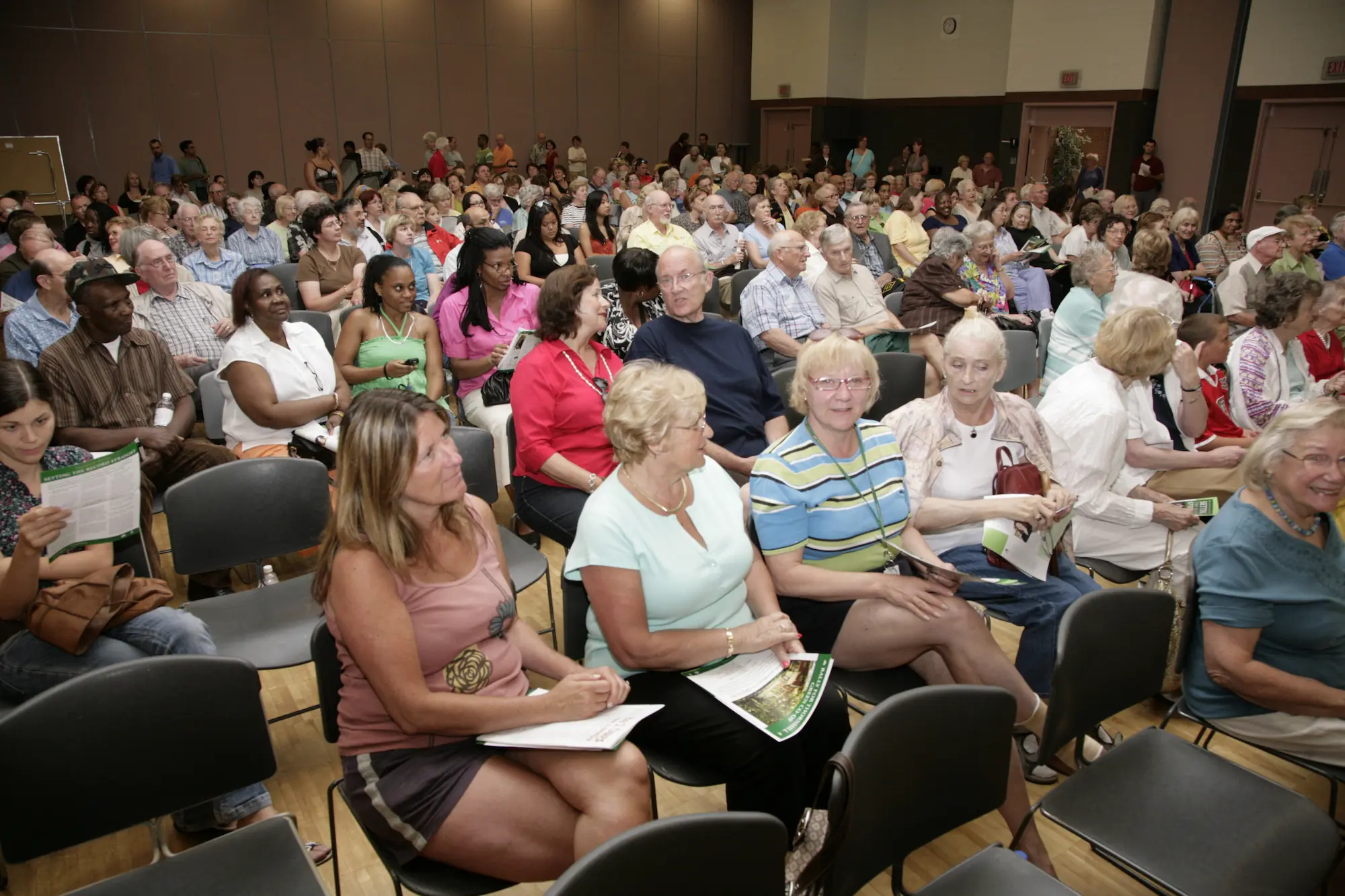 A large group of people sits in rows of chairs inside a crowded auditorium, many holding papers and engaging in conversation before a CHF Canada Co-operative Housing event begins.