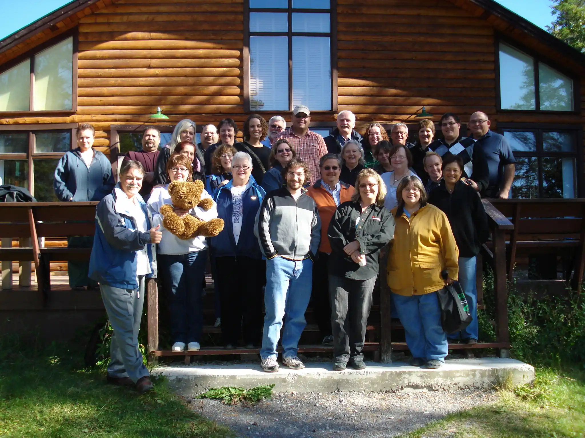 A group of people poses for a photo on the steps of a wooden lodge in daylight; one person in front holds a teddy bear, reflecting the welcoming spirit of co-operative housing and community supported by CHF Canada.