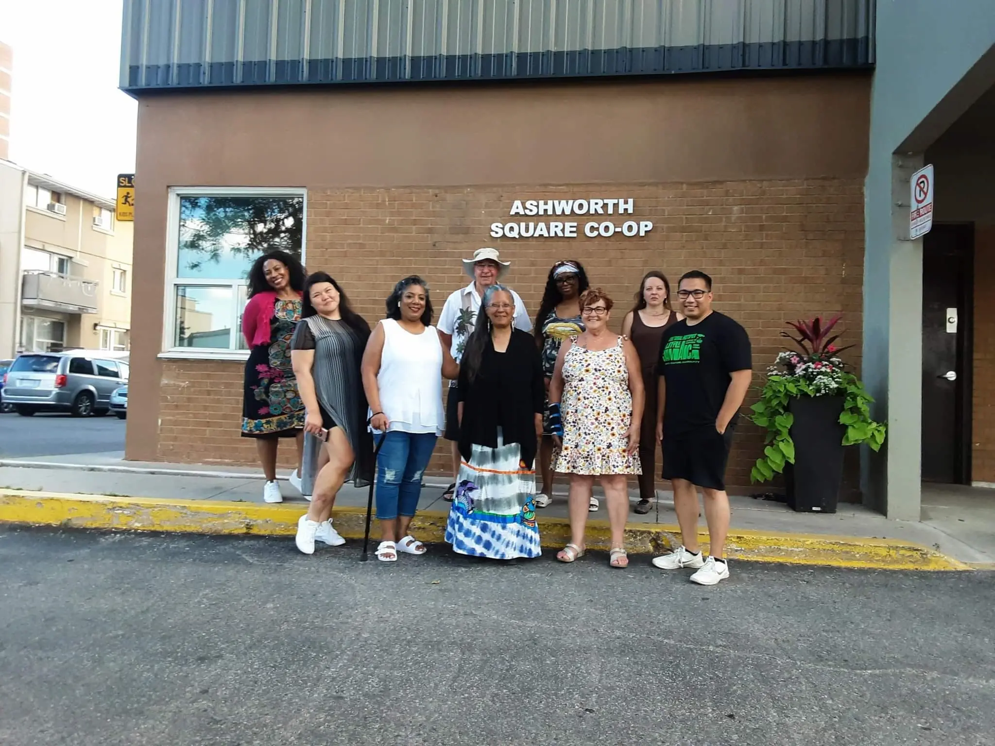 Nine people stand together outside a building labeled "Ashworth Square Co-op," posing for a group photo near the entrance, celebrating the spirit of co-operative housing.