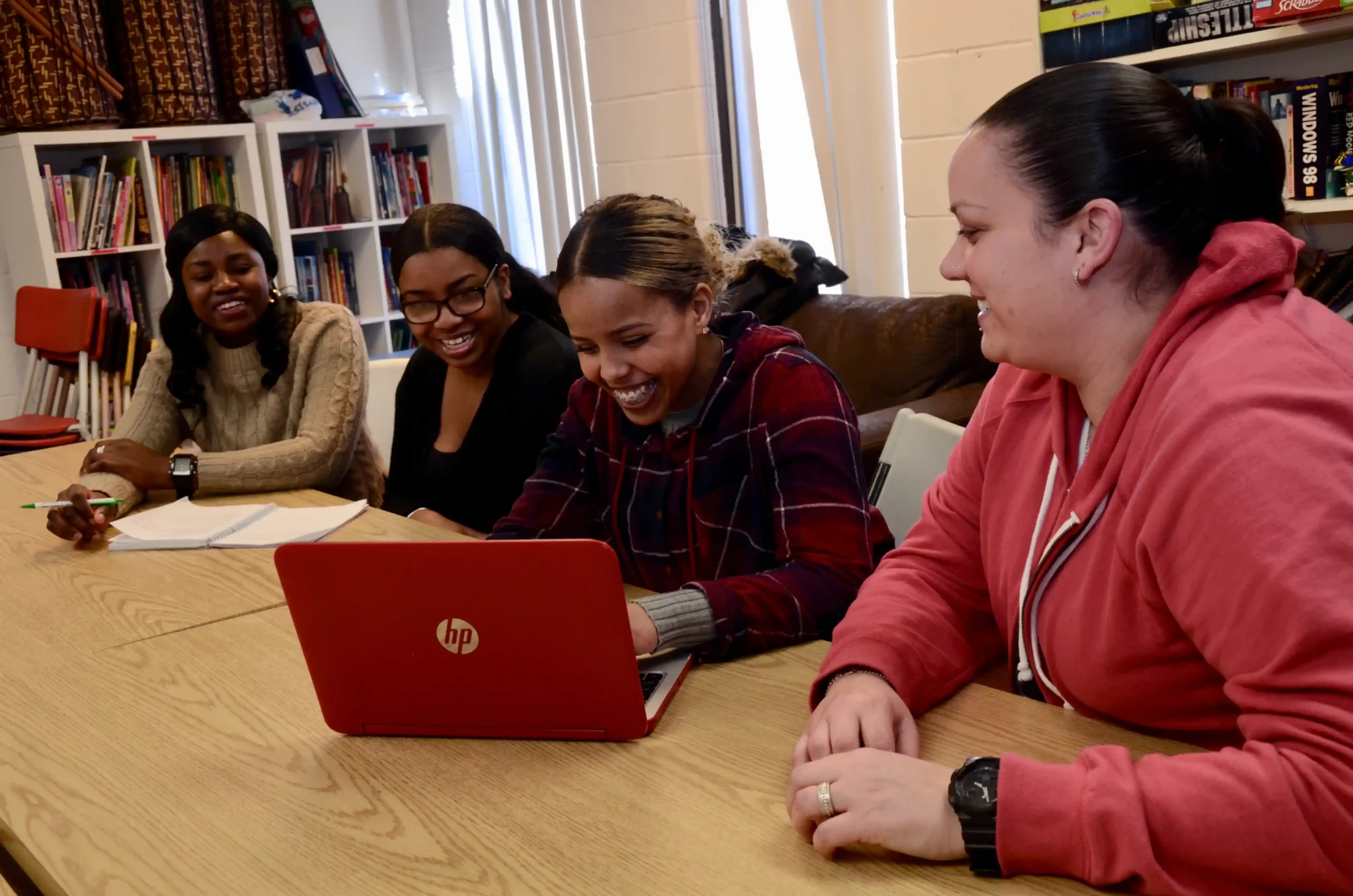 Four women sit at a table with notebooks and a red HP laptop, smiling and engaging in conversation about Co-operative Housing in a room with bookshelves, representing the community spirit of CHF Canada.