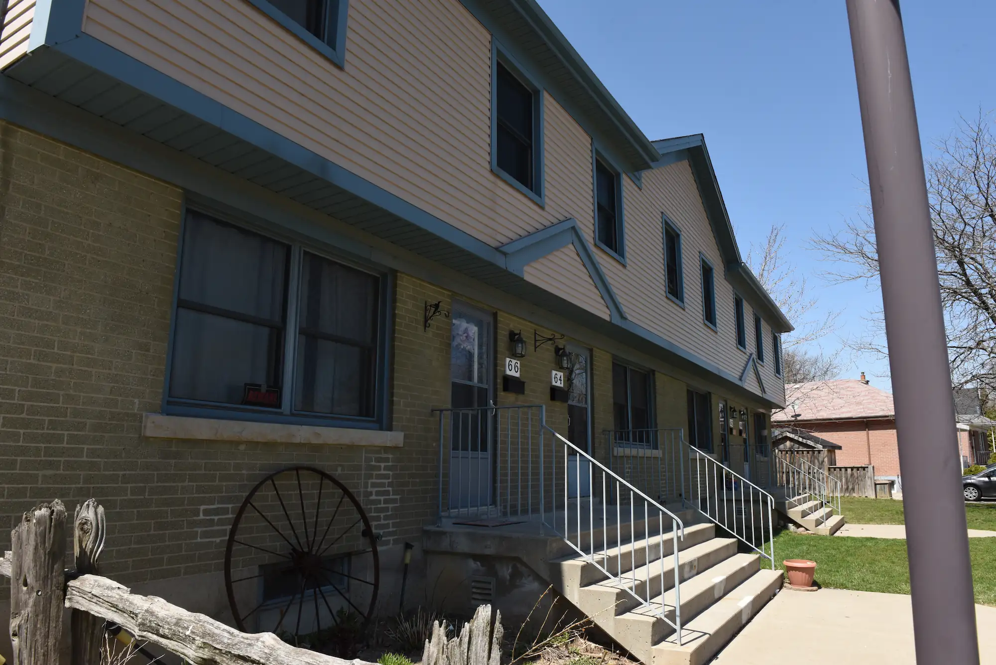 A two-story co-operative housing apartment building with beige brick and siding exterior, metal railings, a set of stairs, and a wagon wheel leaning against a wooden fence.