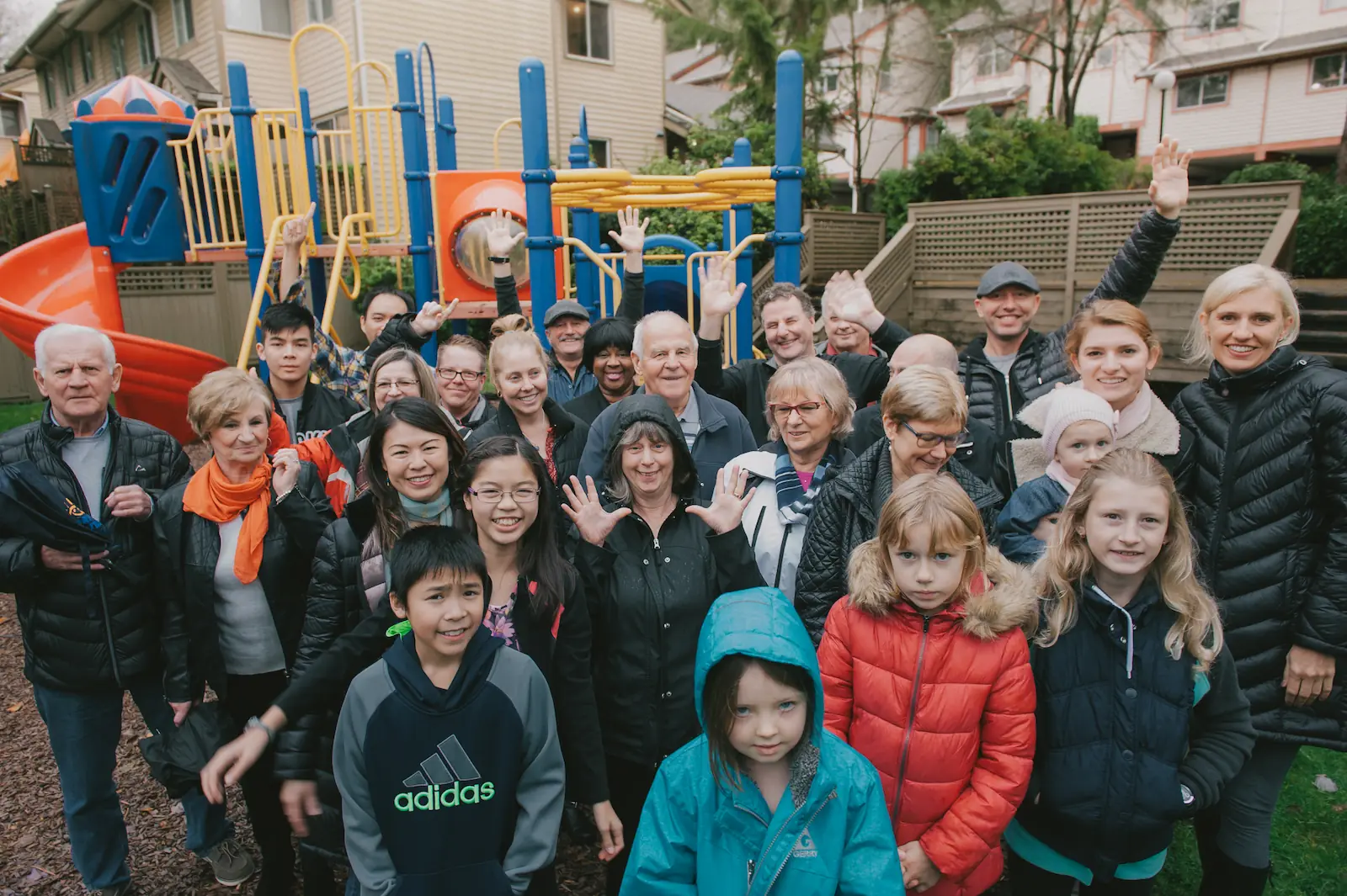 A group of adults and children stand together outdoors in front of a playground structure, posing and smiling at the camera, celebrating their vibrant co-operative housing community.