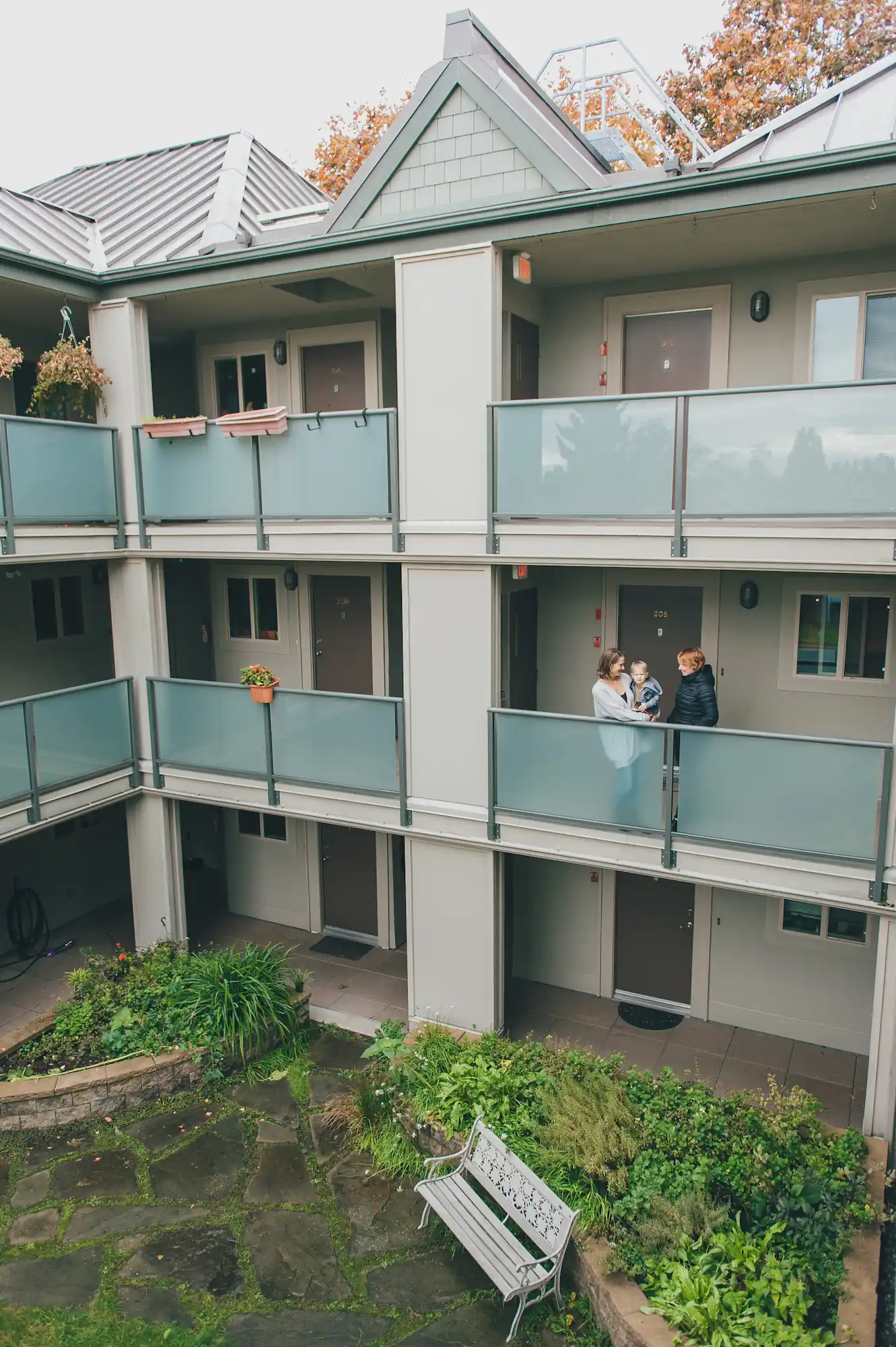 Two people stand and talk on the second-floor balcony of a three-story co-operative housing apartment building, with greenery and a bench in the courtyard below.