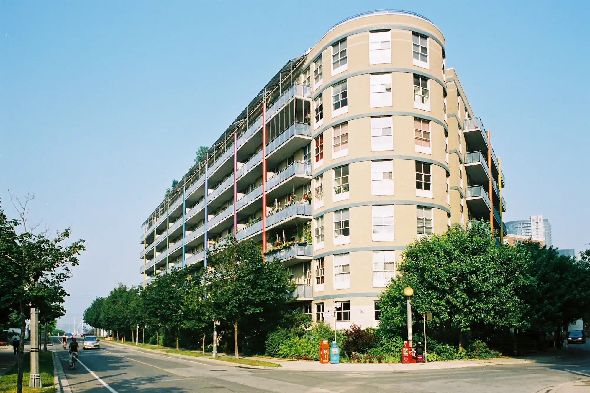 A multi-story co-operative housing apartment building with curved corners and large balconies, surrounded by trees and located at a street intersection on a clear day.