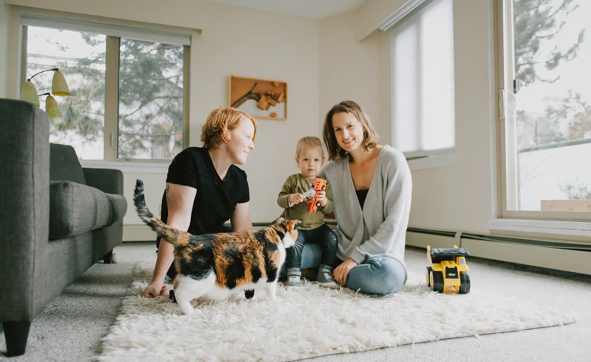 Three people and a toddler sit on a living room floor with a cat walking in front of them and a toy truck nearby, capturing the warmth and community spirit often found in Co-operative Housing supported by CHF Canada.