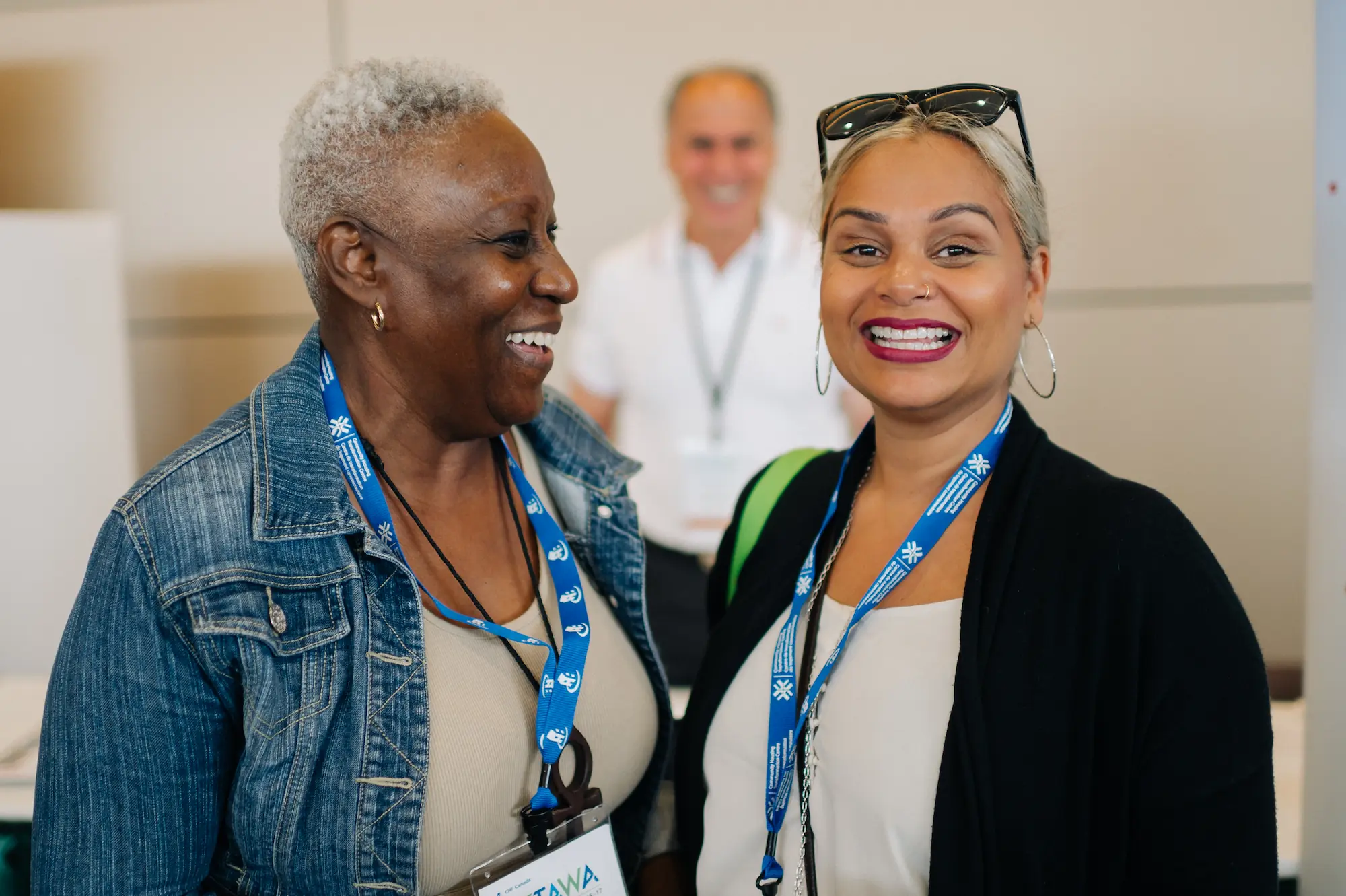 Two women wearing lanyards smile and stand together indoors, with a smiling man in the blurred background at a CHF Canada co-operative housing event.