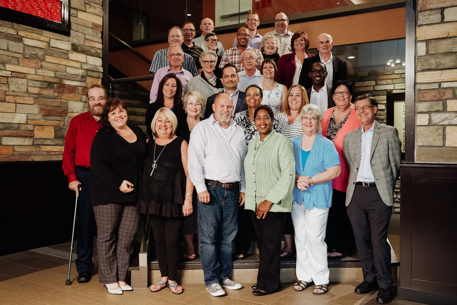 A group of adults posing together on indoor steps, smiling at the camera in a well-lit area with stone walls and glass doors, celebrating the spirit of co-operative housing and community supported by CHF Canada.