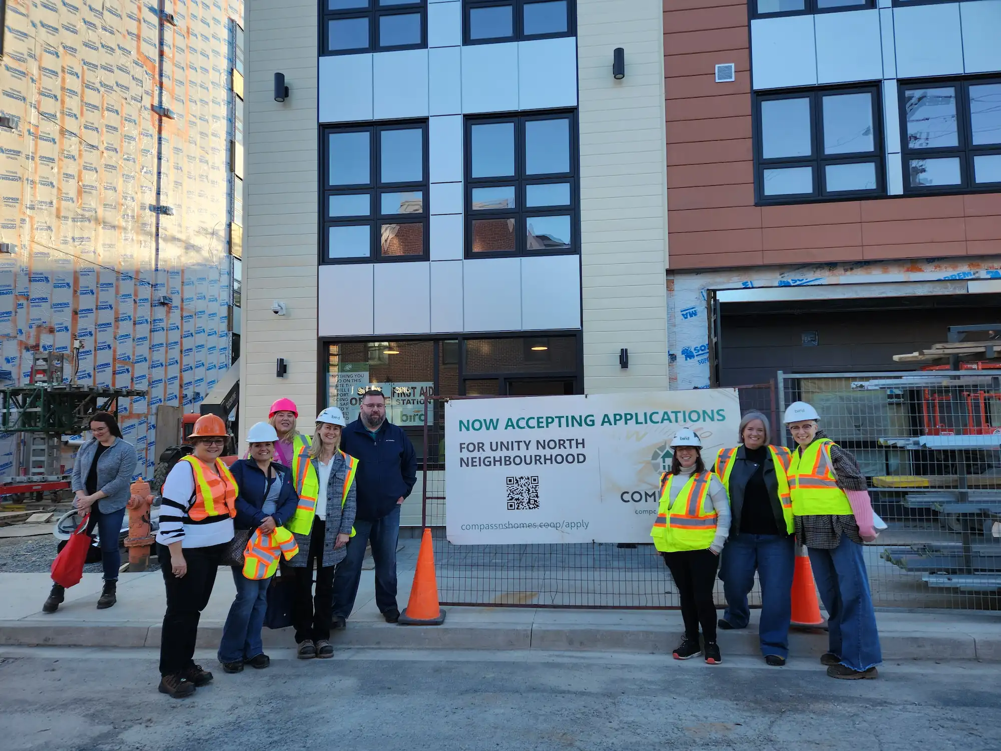 A group of people in safety vests and hard hats stand in front of a building under construction with a sign that reads "Now Accepting Applications for Unity North Neighbourhood Co-operative Housing.